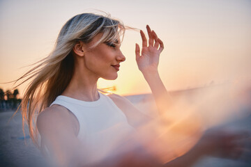 A backlit blonde woman adjusts her hair on a beach with the beautiful gradient of sunset behind her