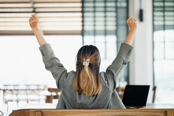 View from behind of a businesswoman with raised arms in victory, celebrating success at her desk with a laptop.