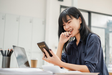 Smiling businesswoman using a smartphone while working on her laptop at a desk with a coffee cup...
