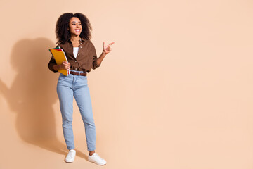 Full size photo of cheerful woman dressed brown shirt hold copybook look directing at sale empty space isolated on beige color background