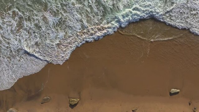 AERIAL TOP DOWN: Ocean waves gently lapping along an empty stretch of sandy beach. Stunning patterns formed by the waves spilling over wet sandy shoreline. Beautiful Atlantic coastline in Portugal.