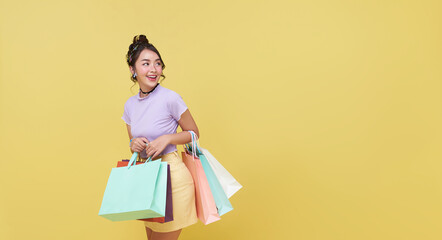 Happy pretty Asian woman carrying colorful shopping bags looking away isolated on yellow studio...