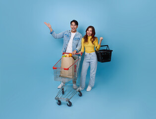 young cheerful Asian couple have fun drive shopping trolley cart and holding shopping bag shopping grocery in supermarket isolated on blue background