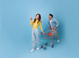 young cheerful Asian couple have fun drive shopping trolley cart and pointing finger in supermarket isolated on blue background