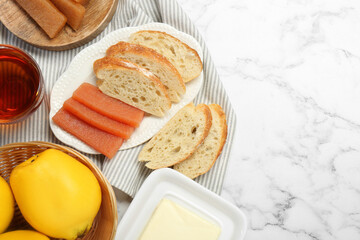 Delicious quince paste, bread, butter, cup of tea and fresh fruits on white marble table, flat lay. Space for text