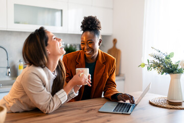 Two female friends laughing while having coffee and working on laptop at home.