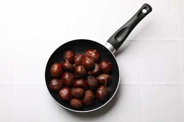 Fresh edible sweet chestnuts in frying pan on white tiled table, top view