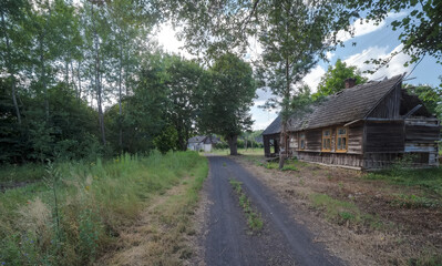 Droga przez wioskę w lesie w letnie popołudnie. Po prawej stronie zabytkowy drewniany dom, który powoli ulega entropii. Powyżej błękitne niebo nad Czarną Gliną. - obrazy, fototapety, plakaty