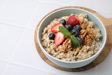 Tasty oatmeal with strawberries, blueberries and walnuts in bowl on white tiled table, closeup. Space for text