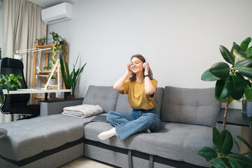 Young woman listening to music with headphones on the couch surrounded by indoor plants. Beautiful girl relaxing at home and listening to a podcast in a wireless headset.