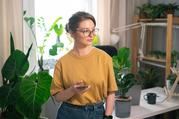 Beautiful young woman working on phone at workstation from home surrounded by indoor plants. Concept of remote work, freelancing, online learning, in the urban jungle.