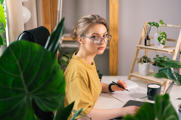 Beautiful young woman working on laptop from home surrounded by indoor plants. Concept of remote work, freelancing, online learning, in the urban jungle.