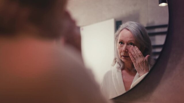 Senior woman touching face skin in front of mirror, displeased with wrinkles