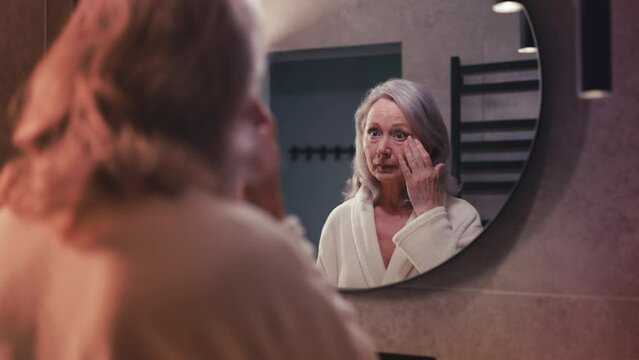 Woman in her 60s looking in the mirror, upset with puffy eyes and dark circles