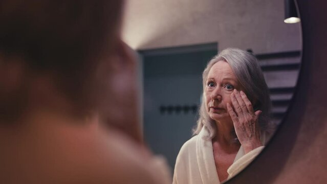 Unhappy senior lady displeased with her skin and wrinkles, looking in mirror