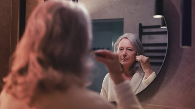 Senior woman using face jade roller for massage, looking in the mirror, skincare