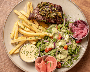 Fotografía corte de carne a la parilla acompañadas de papas fritas y salteadas, con una ensalada verde y aderezo