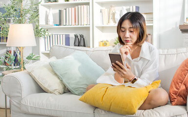 Asian cheerful woman wearing casual clothes, happily smiling, online chatting, using mobile phone, sitting on sofa in indoor living room at cozy home, relax. Recreation, Leisure, Lifestyle Concept.