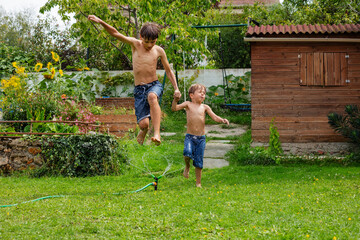 Two kids partake in wet fun, jump over spouts of lawn sprinkler