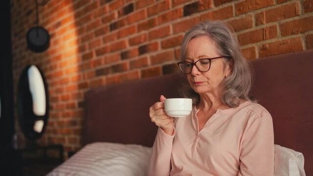Relaxed senior female in glasses drinking morning coffee in bed, inspiration