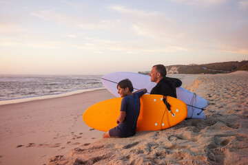 A parent and his son smiling sit with their surfboards on sand