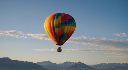 Obraz premium Colorful hot air balloon flying over mountain range, rainbow patterned aerostat in clear blue sky, scenic aerial adventure travel background