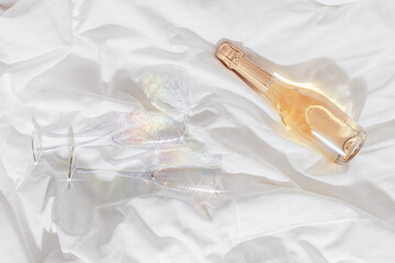 Two colored shiny champagne glasses with white wine bottle on bed, on white sheets. Minimal...