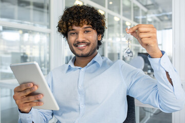 A cheerful young man showcases house keys while using a tablet in a light-filled office. Perfect image conveying success in real estate and technology.