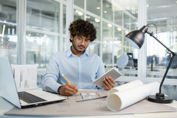 Focused young male architect with curly hair working on architectural plans in a bright, modern office with digital tablet and laptop.