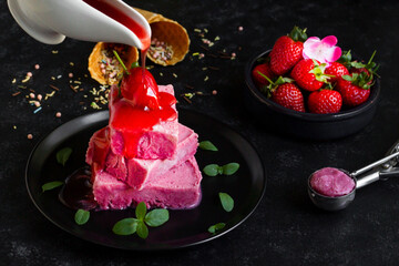 Strawberry ice cream, designed on a black round plate, strawberry sauce pouring from the sauce bowl
