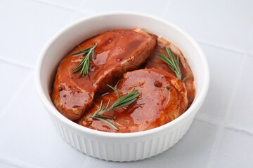 Raw marinated meat and rosemary in bowl on white tiled table, closeup