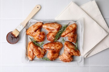 Raw chicken wings, rosemary, marinade and basting brush on light tiled table, flat lay