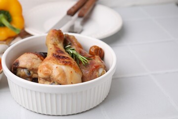 Delicious roasted chicken drumsticks with rosemary and tomatoes in bowl on white tiled table. Space for text