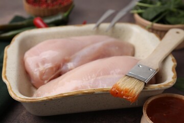 Marinade, basting brush and raw chicken fillets on brown table, closeup