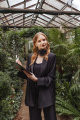 Elegant caucasian woman in black evening jumpsuit uses a stylus with a tablet in a lush garden....