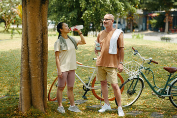 Active mature diverse couple standing under tree and drinking water after bicycle ride