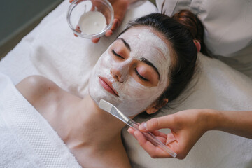 Therapist's hands applying a face mask to a beautiful young man in a spa using a cosmetics brush. Beauty treatment, skin care, wellbeing. - obrazy, fototapety, plakaty