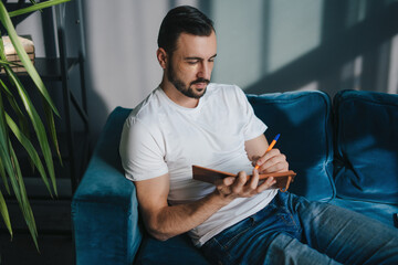 Bearded man looking down thoughtfully while writing in his journal at home. Relaxing time at home.