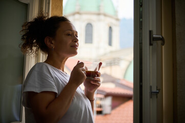Delightful young woman enjoying her morning coffee, standing at the window overlooking the medieval cathedral of Santa Maria di Assunta in the Italian City of Como. - obrazy, fototapety, plakaty