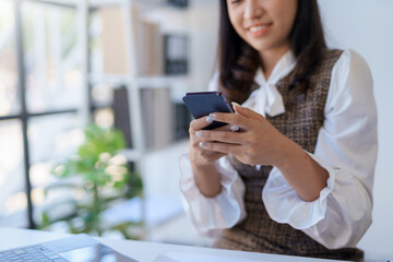 A woman in a white blouse and brown skirt is sitting at a desk in an office, looking at her phone.