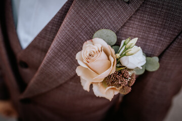 Valmiera, Latvia- July 28, 2024 - A close-up of a boutonniere with a peach rose and green accents pinned on a brown textured suit jacket, part of a formal wedding attire. - obrazy, fototapety, plakaty