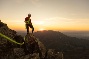 A man in a red jacket is standing on a rock with a green rope attached to him. The sun is setting...