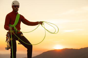 A man in a red jacket is holding a green rope. The rope is attached to a harness on his back. The...