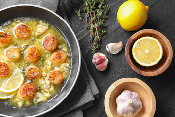 Delicious scallops with sauce in frying pan and ingredients on dark gray textured table, flat lay