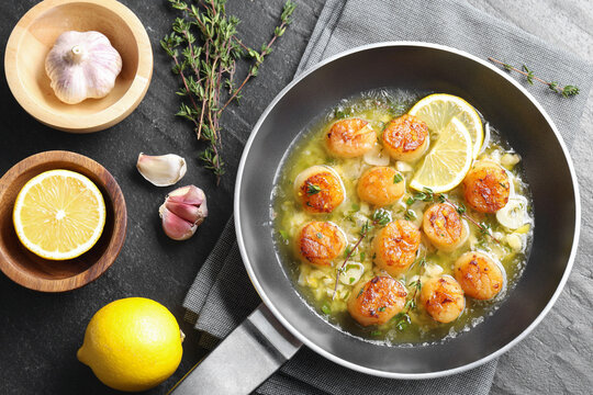 Delicious scallops with sauce in frying pan and ingredients on dark gray textured table, flat lay