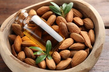Bowl with almond oil in bottle, leaves and nuts on wooden table, closeup