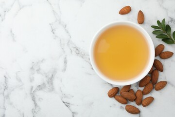 Almond oil in bowl, leaves and nuts on white marble table, flat lay. Space for text