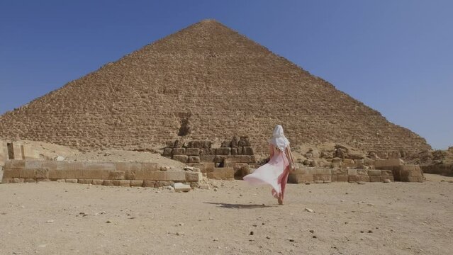 Back view of the young woman walking towards the Great Pyramids of Giza in Egypt and raising hands up. Tourist travel concept. 