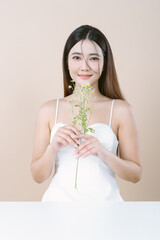 Smiling beautiful Asian woman in white dress holding Gypsophila flower and looking at camera on beige background. Skin care beauty treatments concept. White model with clean, health skin of face.