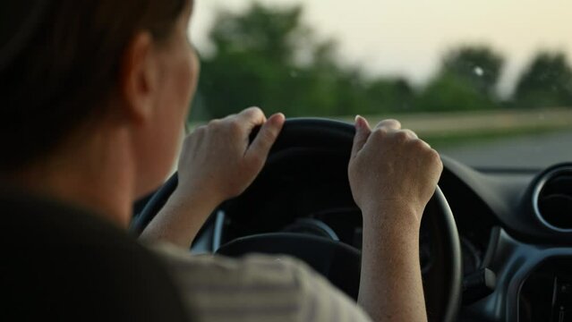 Rear view of female driver holding firmly car steering wheel and driving along the countryside road in summer sunset